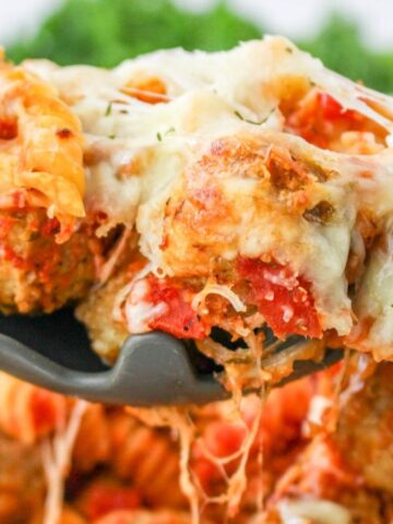 A spoonful of cheesy baked pasta with meatballs, tomato sauce, and melted cheese above a casserole dish.