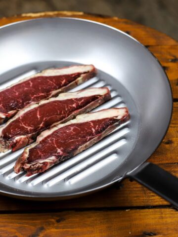 Three raw steak strips in a metal grill pan on a rustic wooden table, ready to be cooked.
