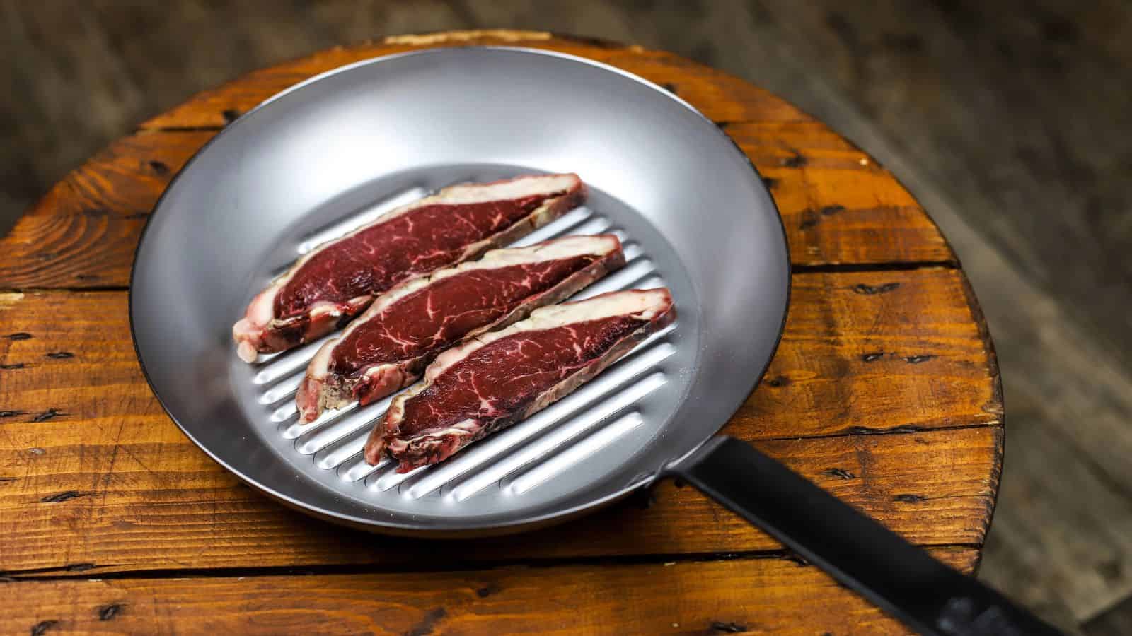 Three raw steak strips in a metal grill pan on a rustic wooden table, ready to be cooked.