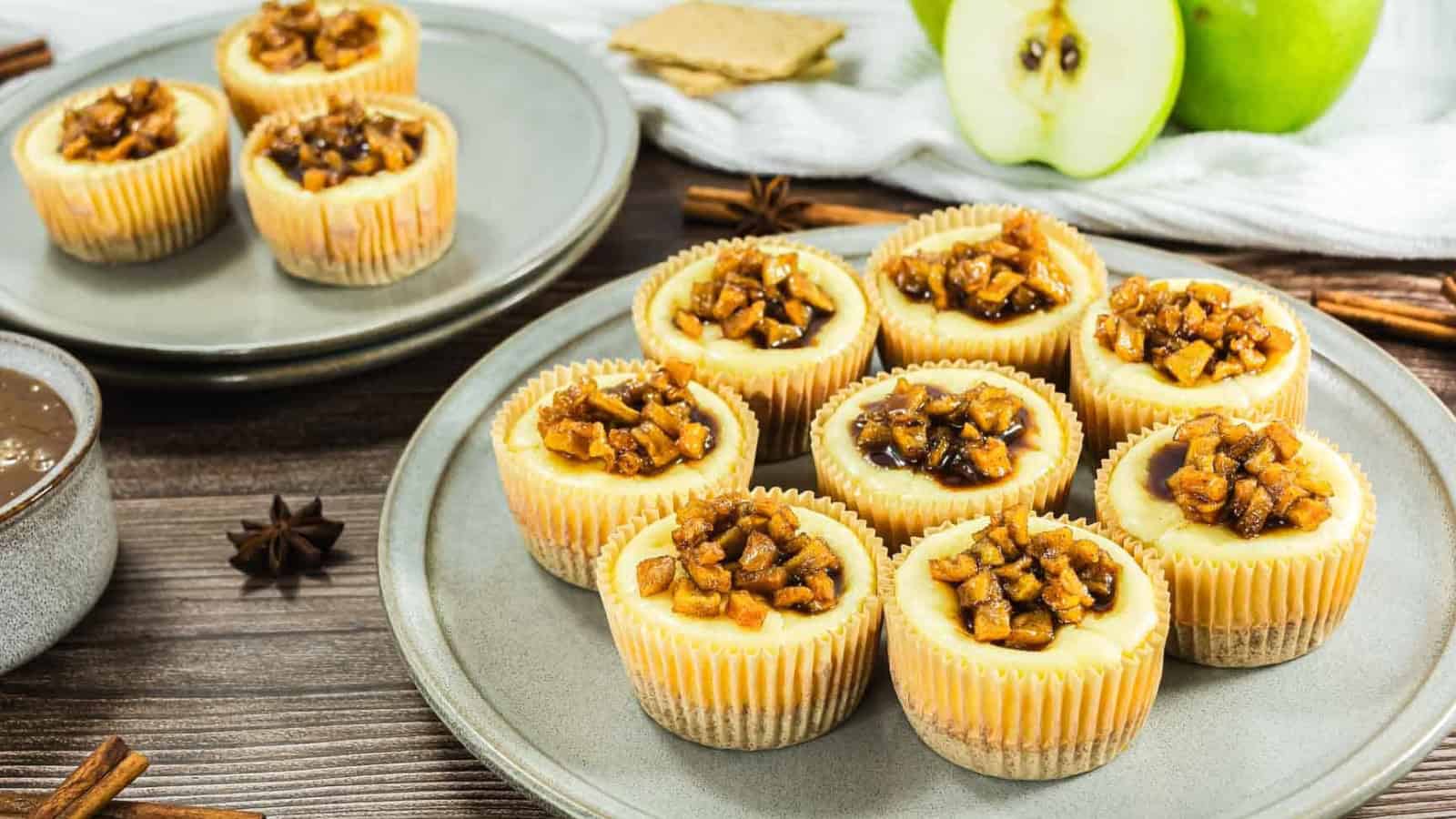Mini cheesecakes topped with caramelized apples on plates, with a green apple and graham crackers in the background.