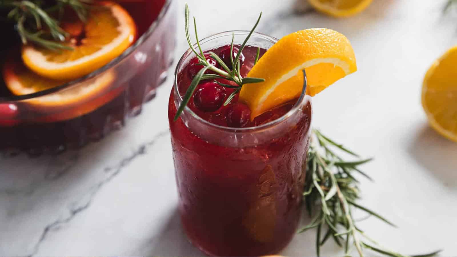 A glass of red punch with ice, cranberries, orange slice, and rosemary garnish on a marble surface.