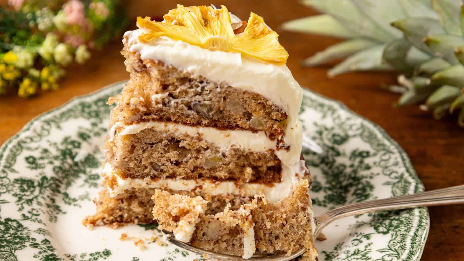 A slice of Hummingbird Cake with cream cheese frosting and pineapple chunks on top, placed on a decorative green and white plate. The Southern cake appears moist with visible nuts, and a fork is resting at the base.