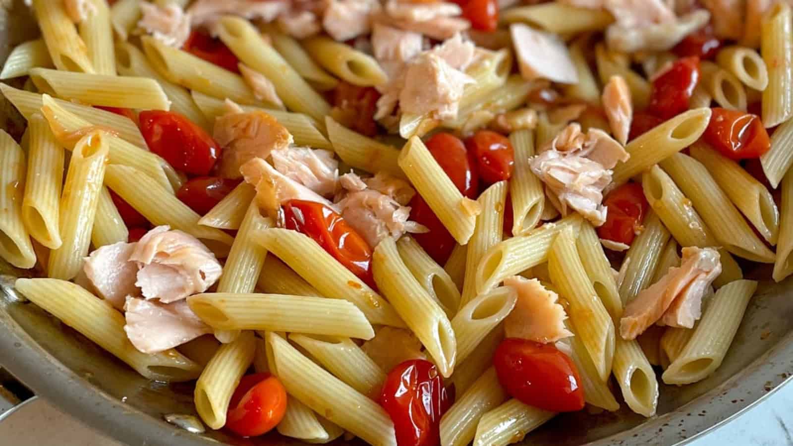 Penne pasta with burst cherry tomatoes, shallots, garlic, and parsley on white plate next to a pan filled with pasta.
