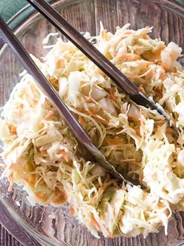 Glass bowl of creamy coleslaw with tongs, on a wooden table next to a green cloth.