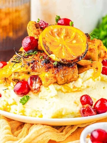 Grilled chicken with lemon, herbs, and cranberries served over mashed potatoes on a white plate.