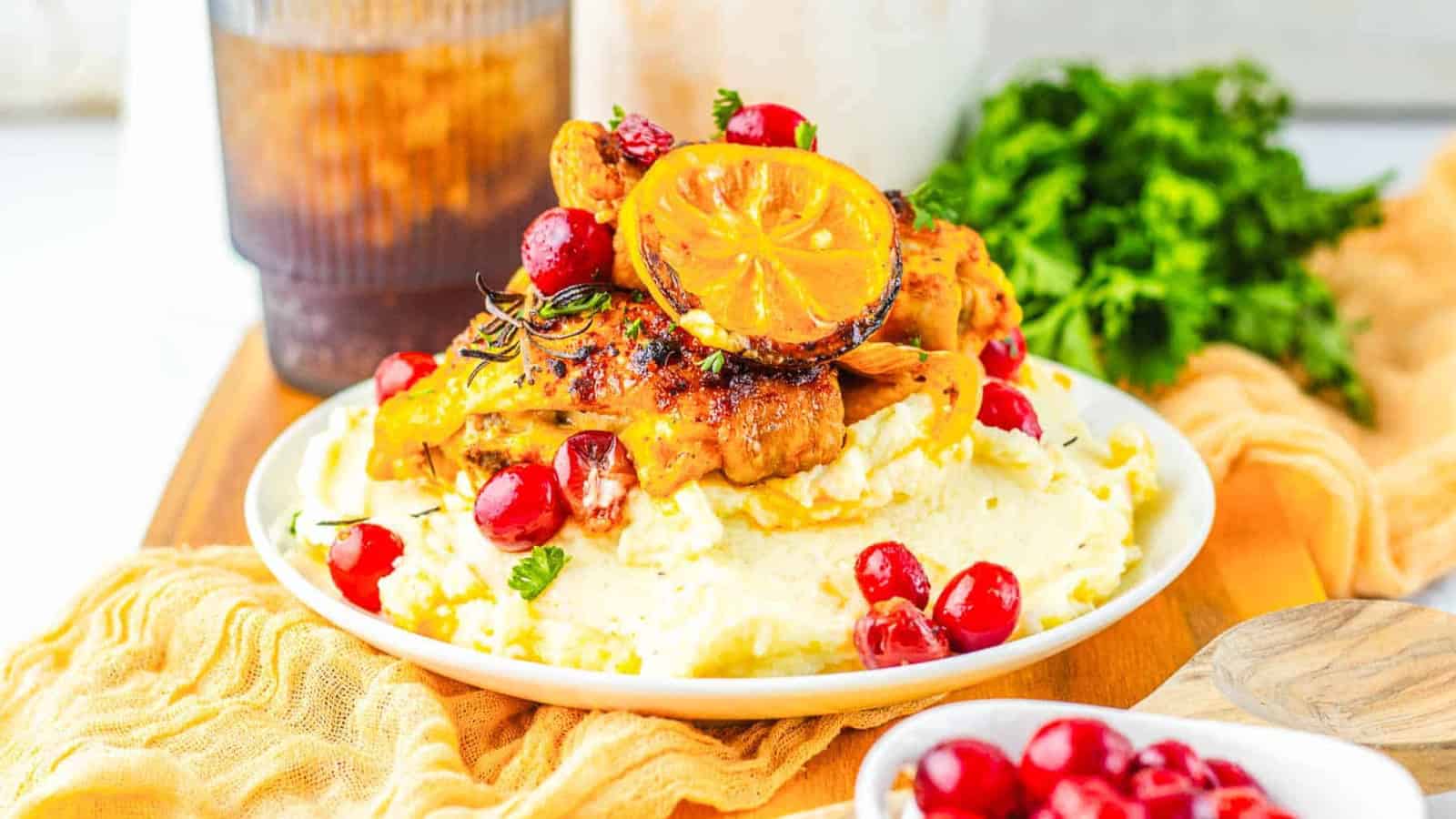 Roasted chicken on mashed potatoes, garnished with cranberries and a grilled lemon slice, with herbs in the background.