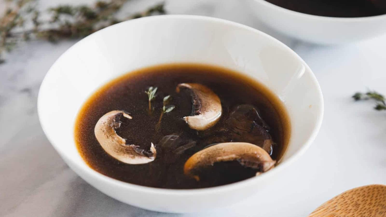 A white bowl of dark mushroom broth with three mushroom slices and a sprig of thyme on top.