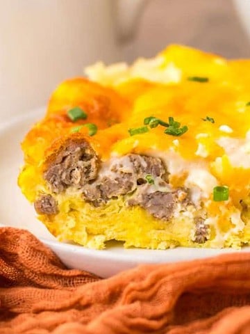 A slice of cheesy breakfast casserole with sausage and chives on a white plate by an orange napkin.