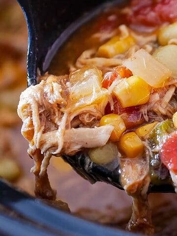 A ladle scooping vegetable stew with corn, tomatoes, beans, and shredded meat from a pot.