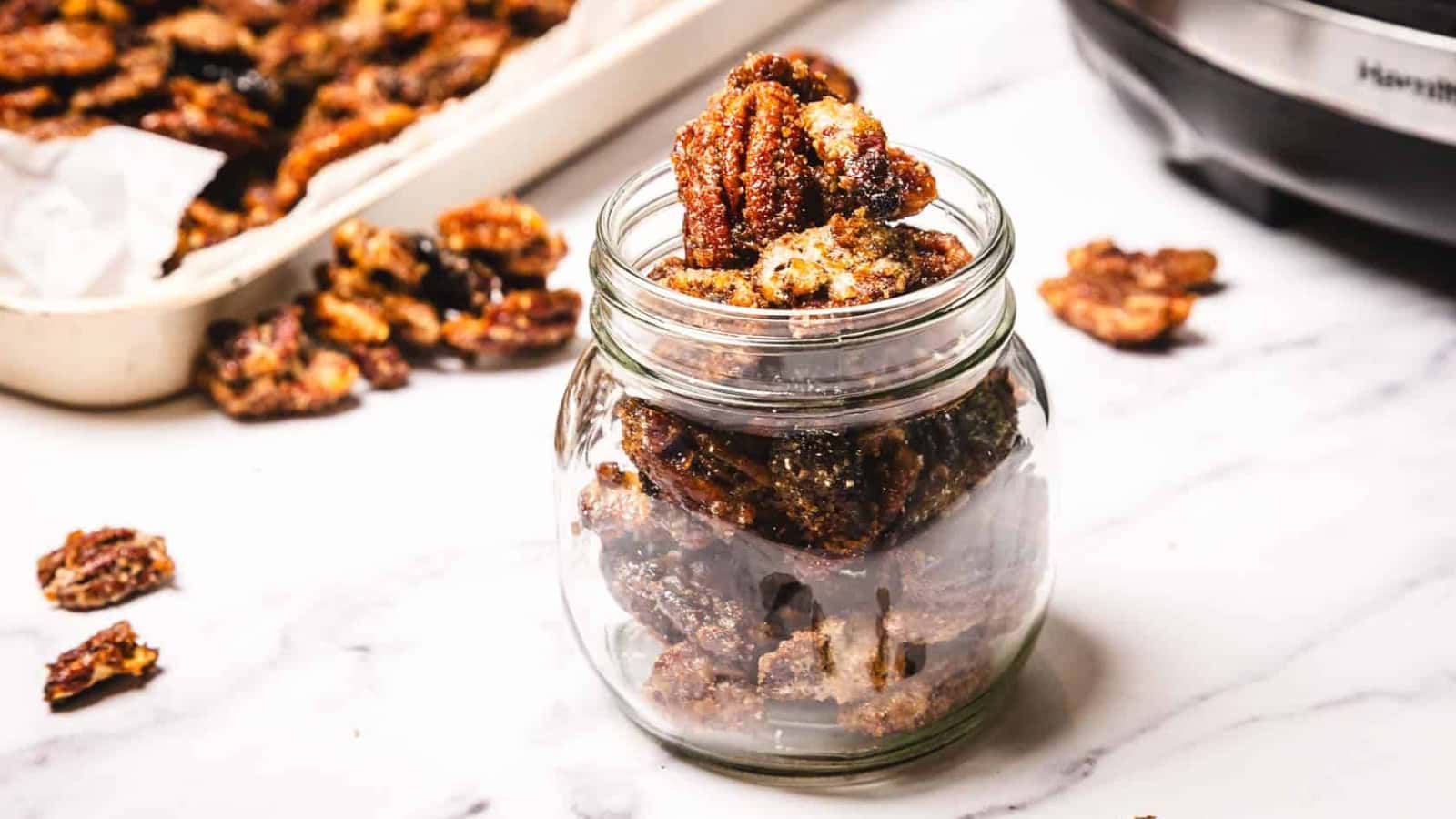 A glass jar filled with candied pecans, with more pecans on a tray and scattered on a white surface nearby.