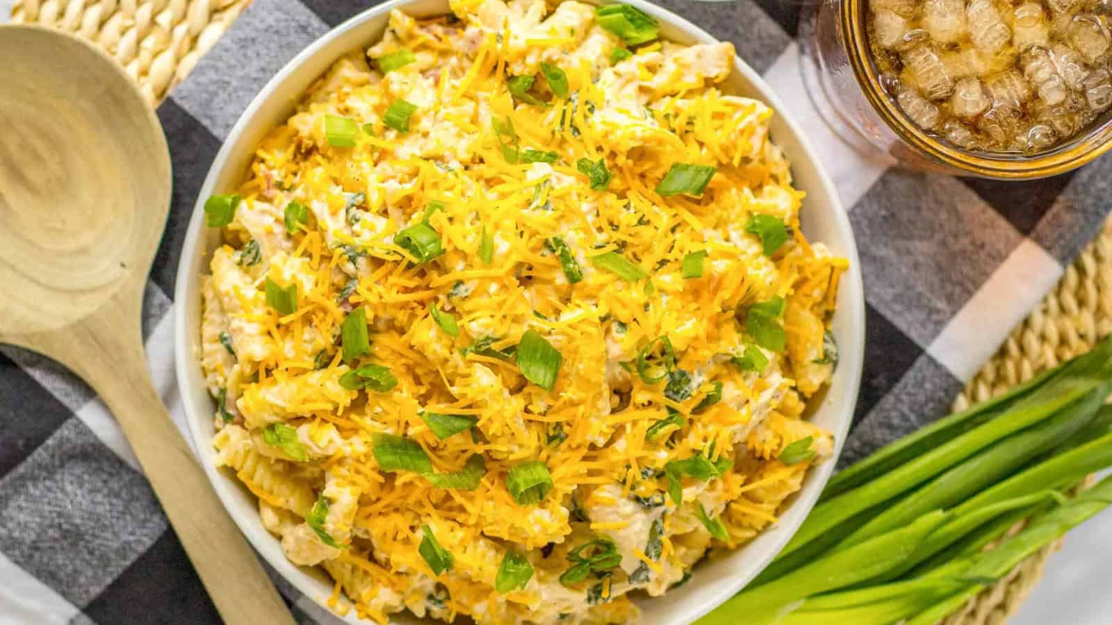 A bowl of creamy rotini pasta salad topped with shredded cheddar cheese and chopped green onions.