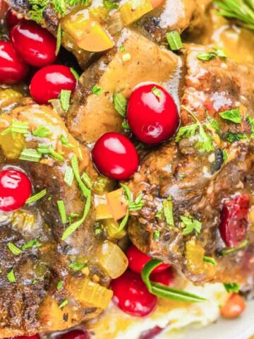 Braised meat with gravy, cranberries, and herbs served over mashed potatoes on a white plate.