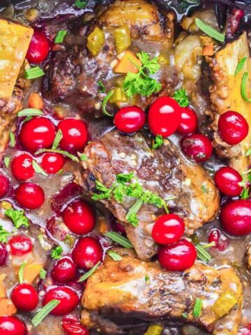Slow-cooked beef stew with red cranberries, vegetables, and fresh herbs in a black crockpot.