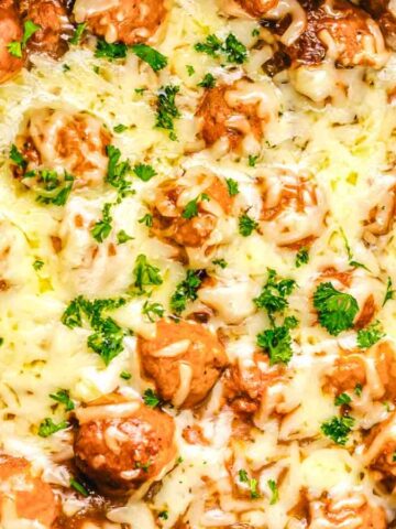 Meatballs in gravy topped with melted cheese and chopped parsley in a black slow cooker.