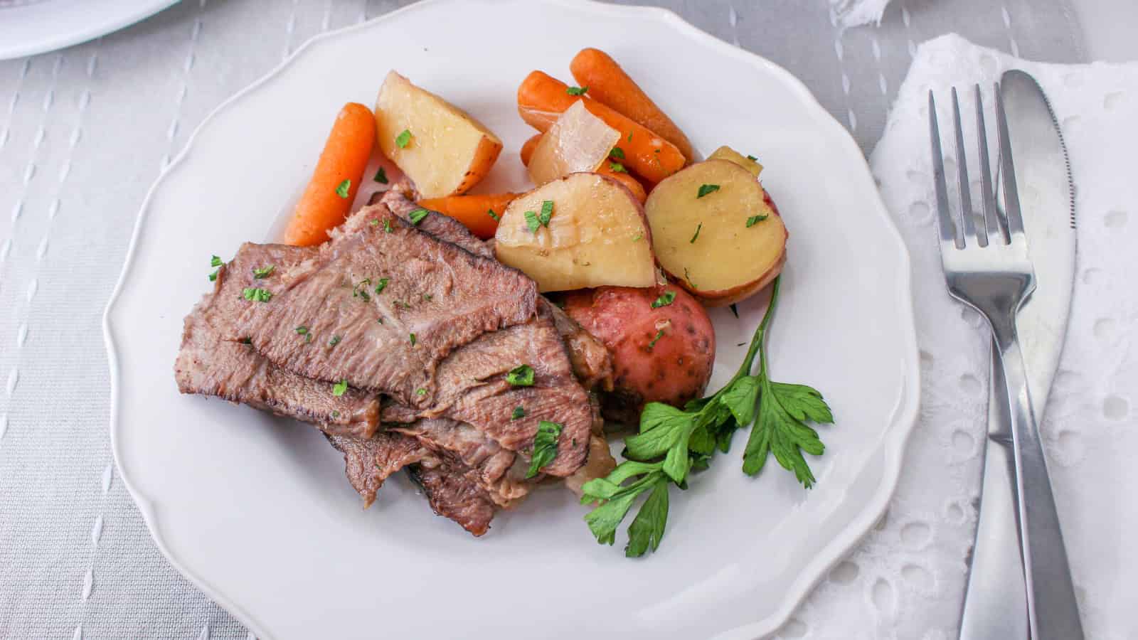 A plate with sliced beef, carrots, potatoes, and parsley garnish, placed beside a fork and knife on a white table setting.
