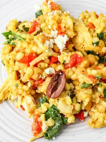 A plate of couscous salad with vegetables, olives, spinach, chicken, and crumbled feta cheese.