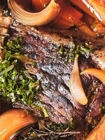 Close-up of a cooked pot roast with carrots, onions, herbs, and rich brown gravy.