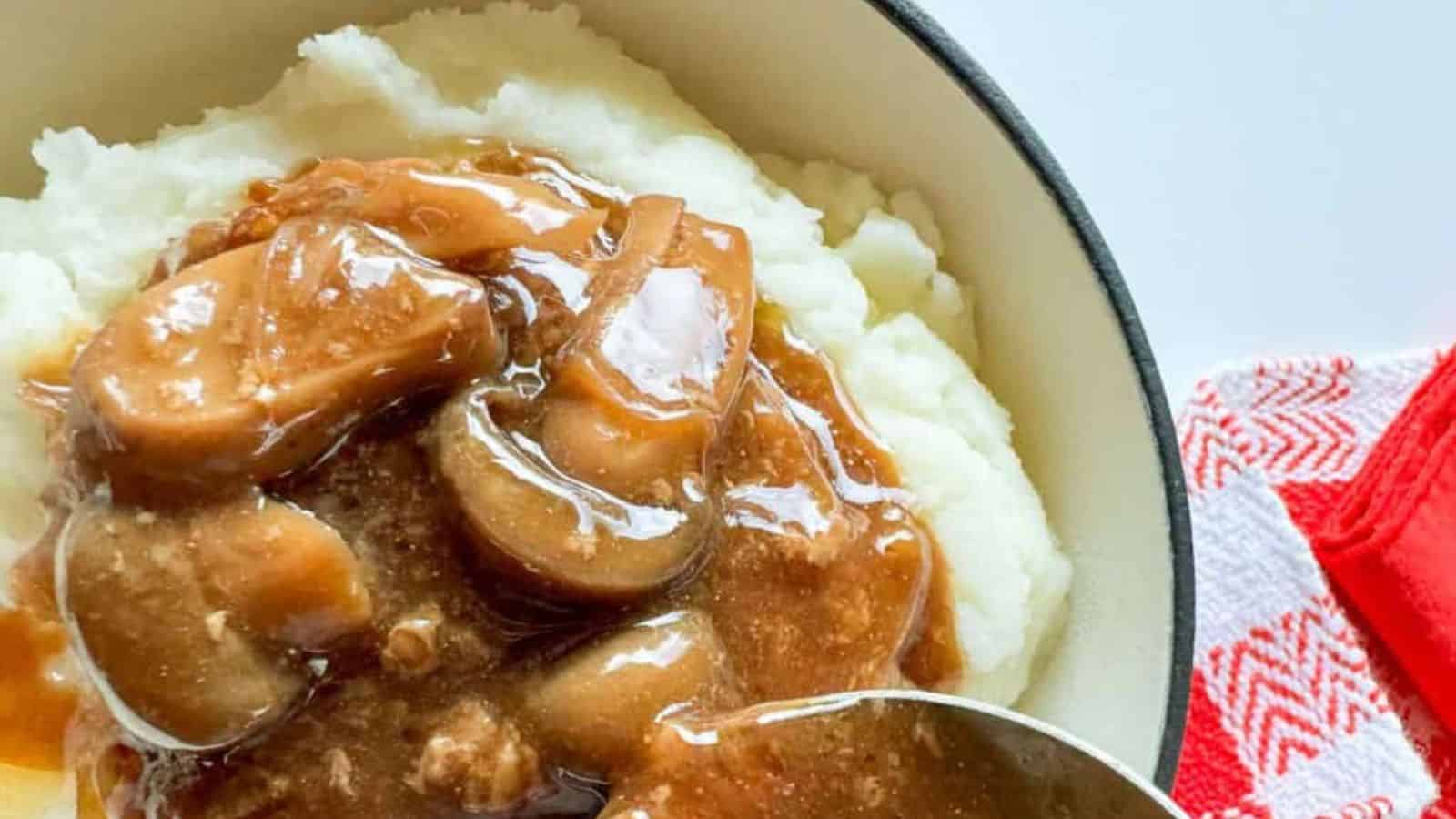 Mashed potatoes topped with brown gravy and mushrooms, reminiscent of a slow cooker Salisbury steak, in a bowl placed on a red and white checkered cloth.