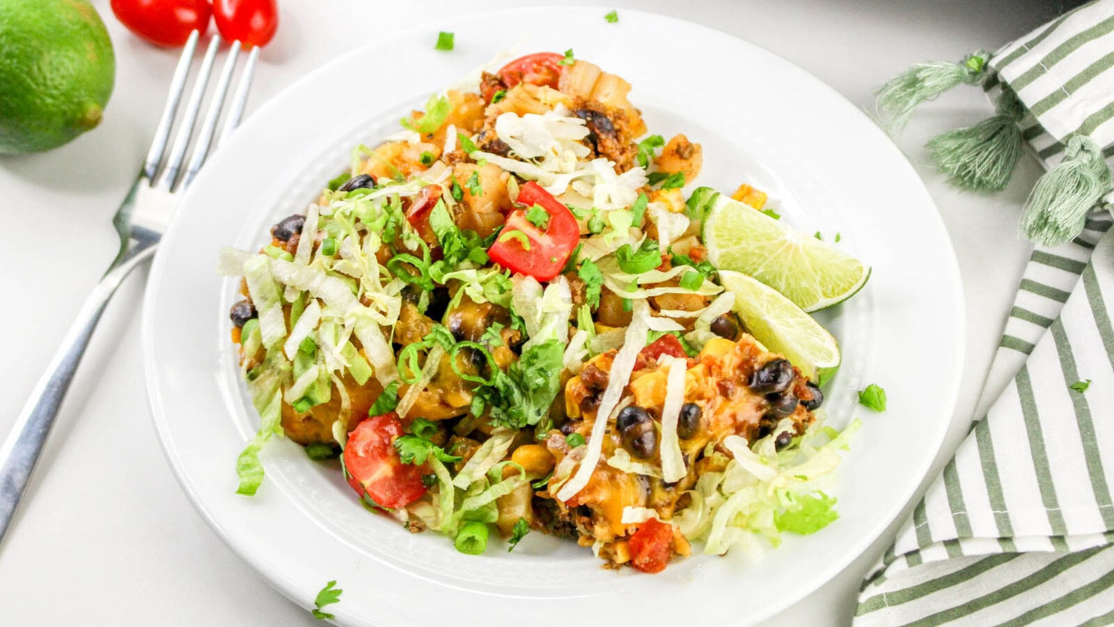Plate of taco casserole with black beans, corn, tomatoes, lettuce, and lime wedges, garnished with shredded cheese and cilantro. Fork and napkin nearby.
