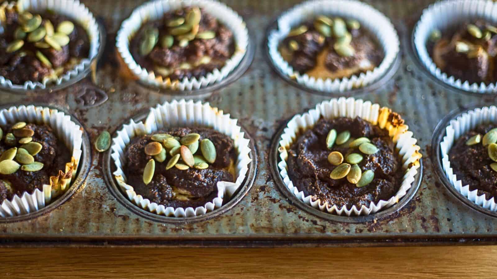 A muffin tin containing eight chocolate muffins lined with white paper cups, each topped with pumpkin seeds. The tin shows signs of use, with some residue and slight rust, and sits on a wooden surface, reminding you that you&rsquo;re missing out on trying new recipes.