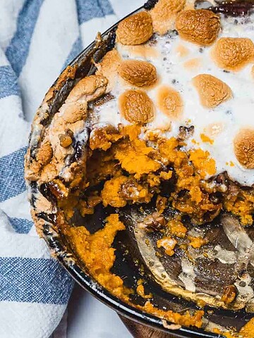 Baked sweet potato casserole topped with toasted marshmallows in a round dish, partially eaten, with a metal serving spoon visible.