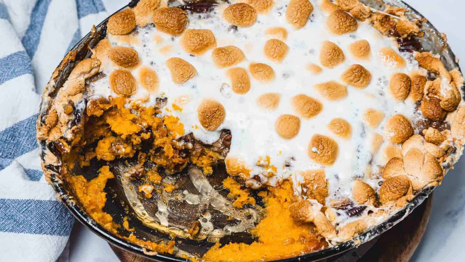 Baked sweet potato casserole topped with toasted marshmallows in a round dish, partially eaten, with a metal serving spoon visible.