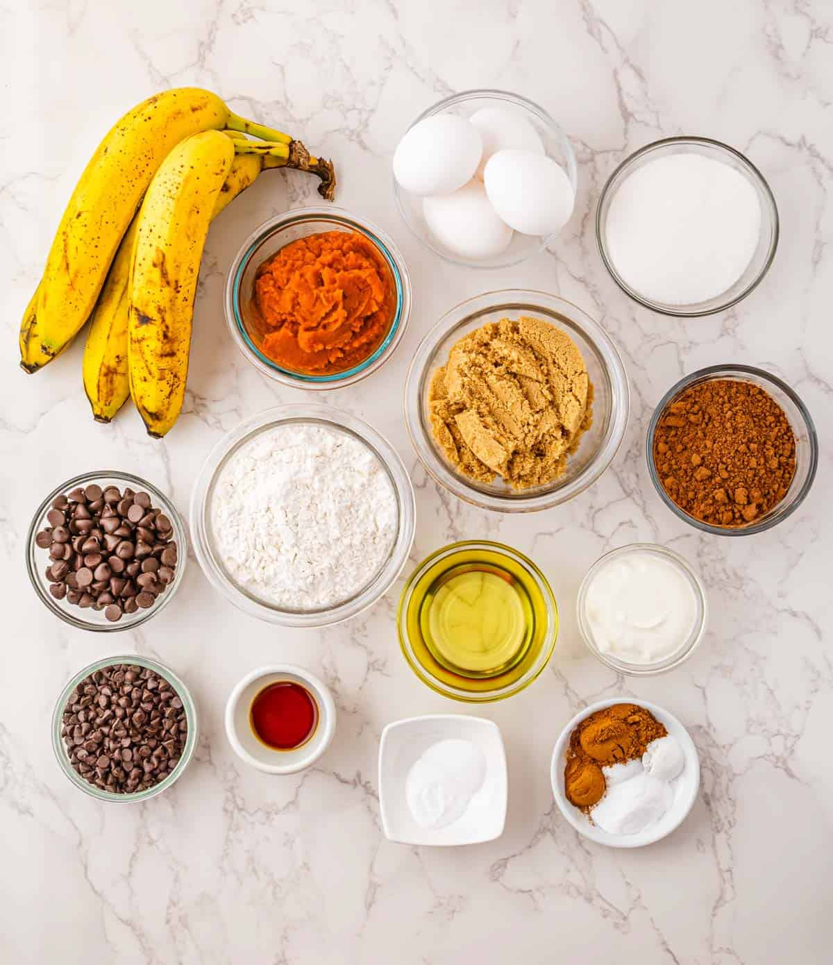 Overhead view of baking ingredients in bowls, including bananas, flour, sugar, eggs, cocoa, oil, and chocolate chips&mdash;perfect for making delicious pumpkin banana bread.