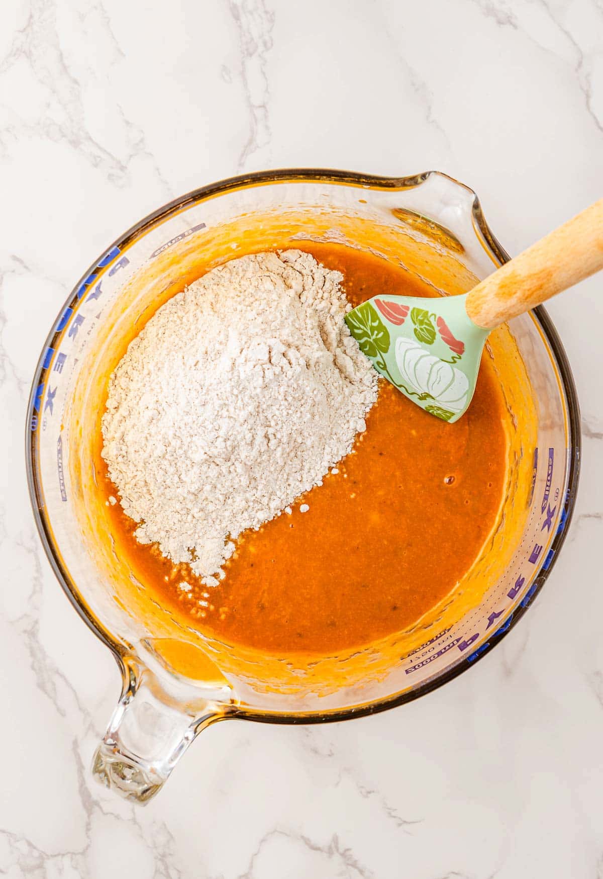Glass bowl with orange batter and flour on top for Pumpkin Banana Bread, being mixed with a green spatula on a marble surface&mdash;perfect for Halloween baking.