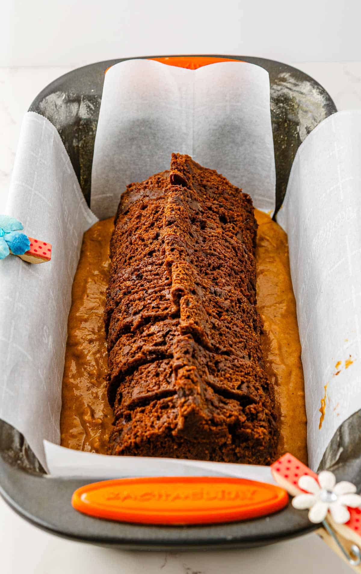 A sliced Pumpkin Banana Bread loaf cake in a parchment-lined pan, with a tan batter underneath&mdash;perfect for Halloween gatherings.