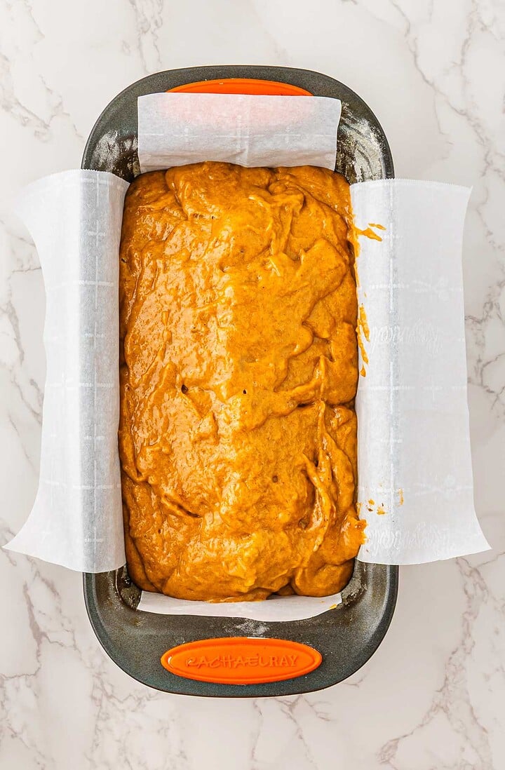 A loaf pan lined with parchment paper filled with Pumpkin Banana Bread batter, ready to be baked for a cozy Halloween treat.