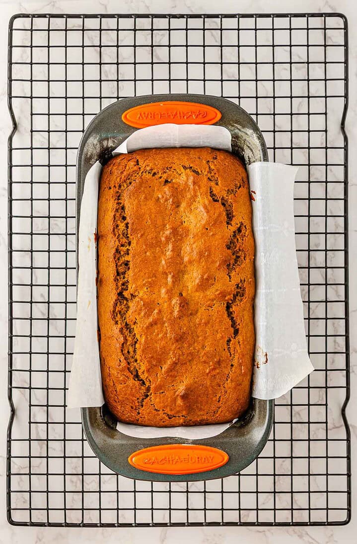 A loaf of Pumpkin Banana Bread in a baking pan lined with parchment paper cools on a rack, perfect for a cozy Halloween treat.