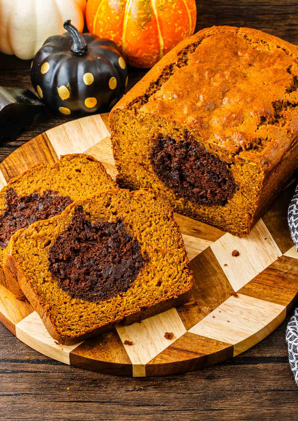 Sliced Pumpkin Banana Bread with a chocolate swirl center, displayed on a wooden board with decorative pumpkins nearby&mdash;perfect for a festive Halloween treat.