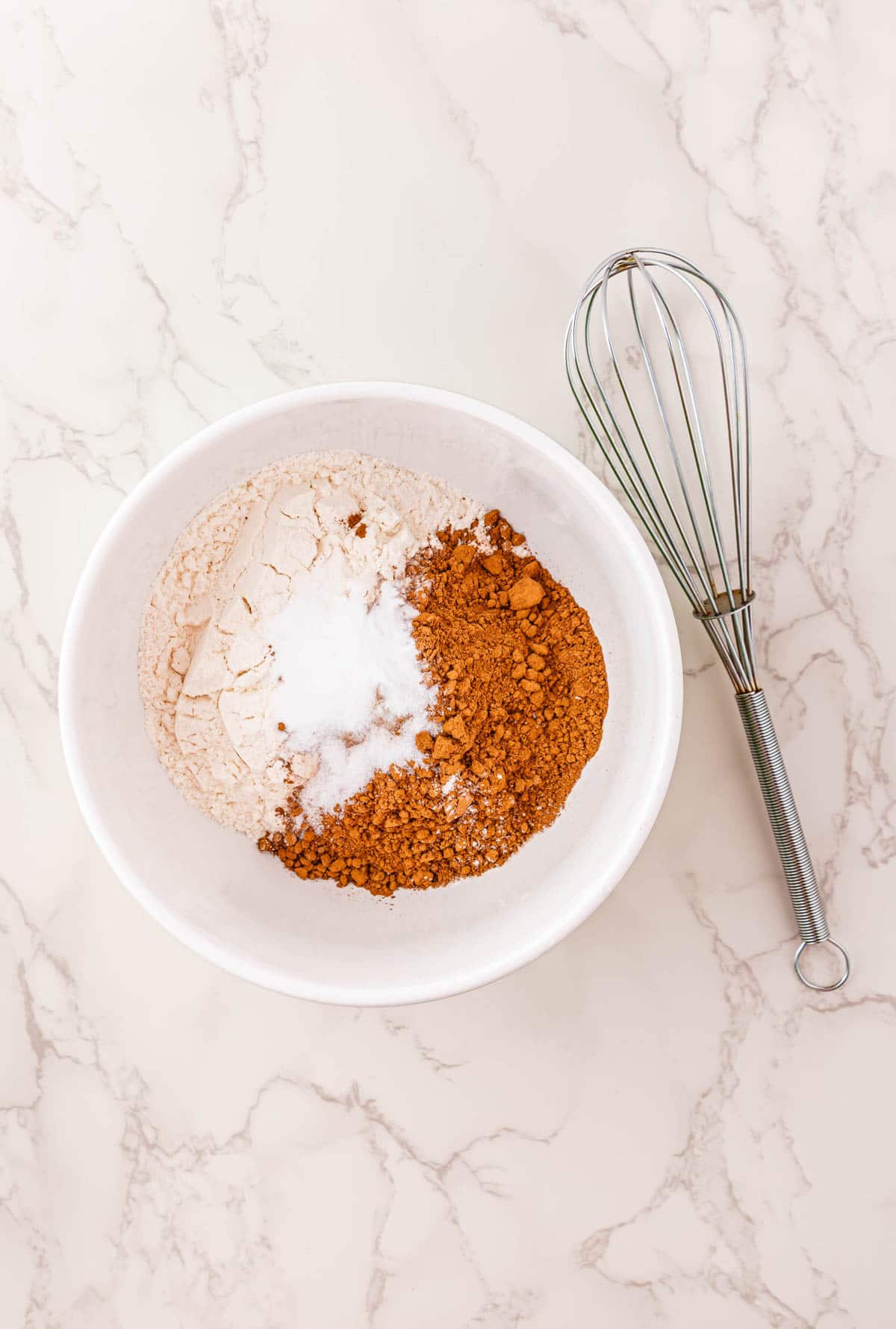 White bowl with flour, cocoa powder, and sugar beside a metal whisk on a marble countertop&mdash;perfect for whipping up Halloween Pumpkin Banana Bread.