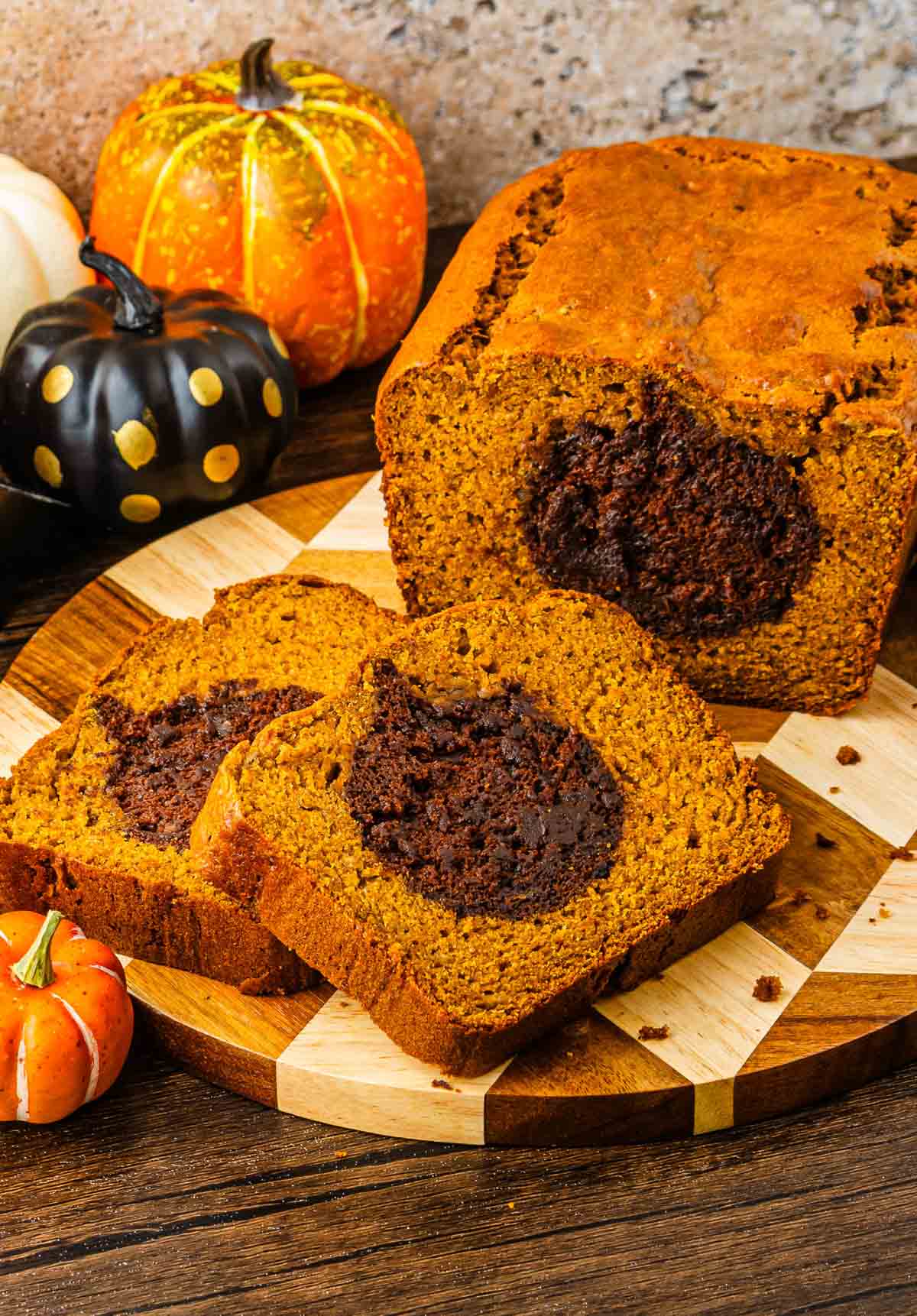 Sliced Pumpkin Banana Bread with a chocolate swirl, displayed on a wooden board alongside decorative pumpkins&mdash;perfect for your Halloween table.
