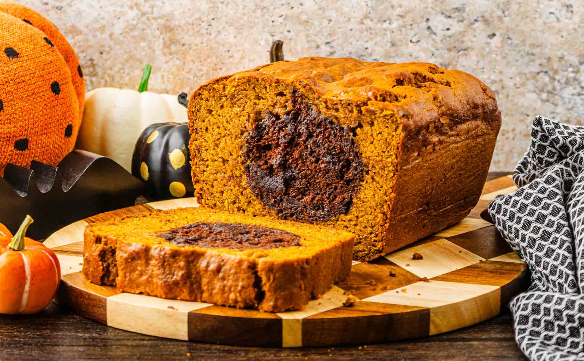 A sliced Pumpkin Bread loaf with a chocolate swirl sits on a wooden board, pumpkins in the background&mdash;a festive Halloween Surprise perfect for autumn gatherings.