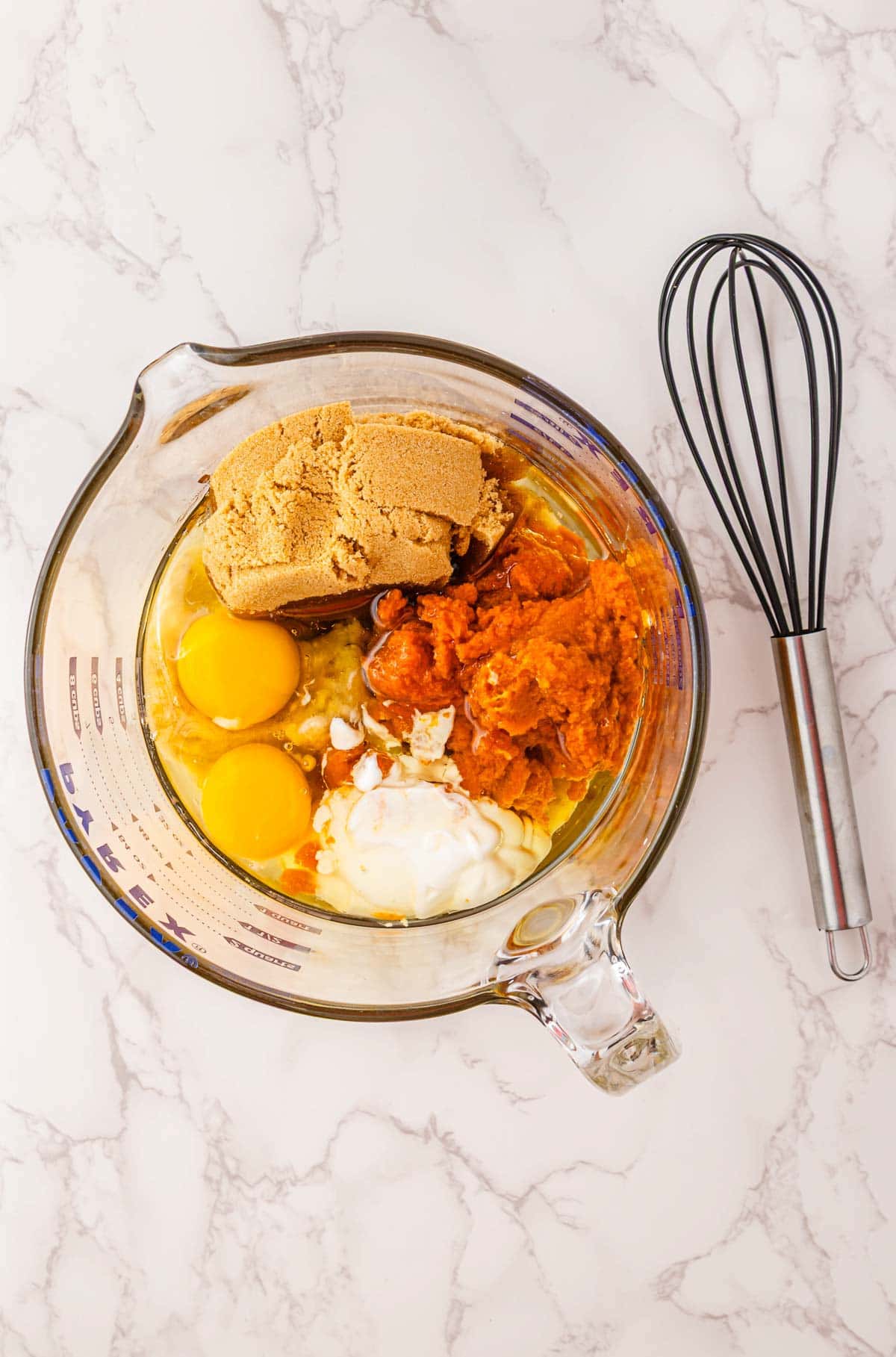 Glass bowl with eggs, brown sugar, pumpkin puree, and other baking ingredients next to a whisk on a marble surface&mdash;perfect for whipping up Pumpkin Banana Bread for Halloween.