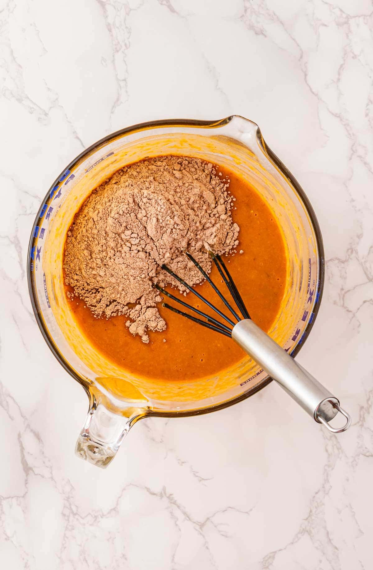 A glass bowl with orange batter and dry cocoa mix, whisk inside, sits on a marble countertop&mdash;perfect for whipping up a Pumpkin Banana Bread surprise this Halloween.