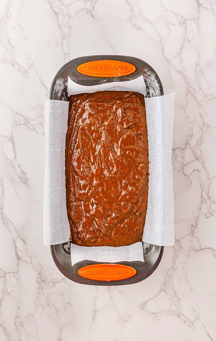 A loaf pan lined with parchment paper, filled with spiced pumpkin banana bread batter, sits on a marble countertop&mdash;perfect for a cozy Halloween treat.