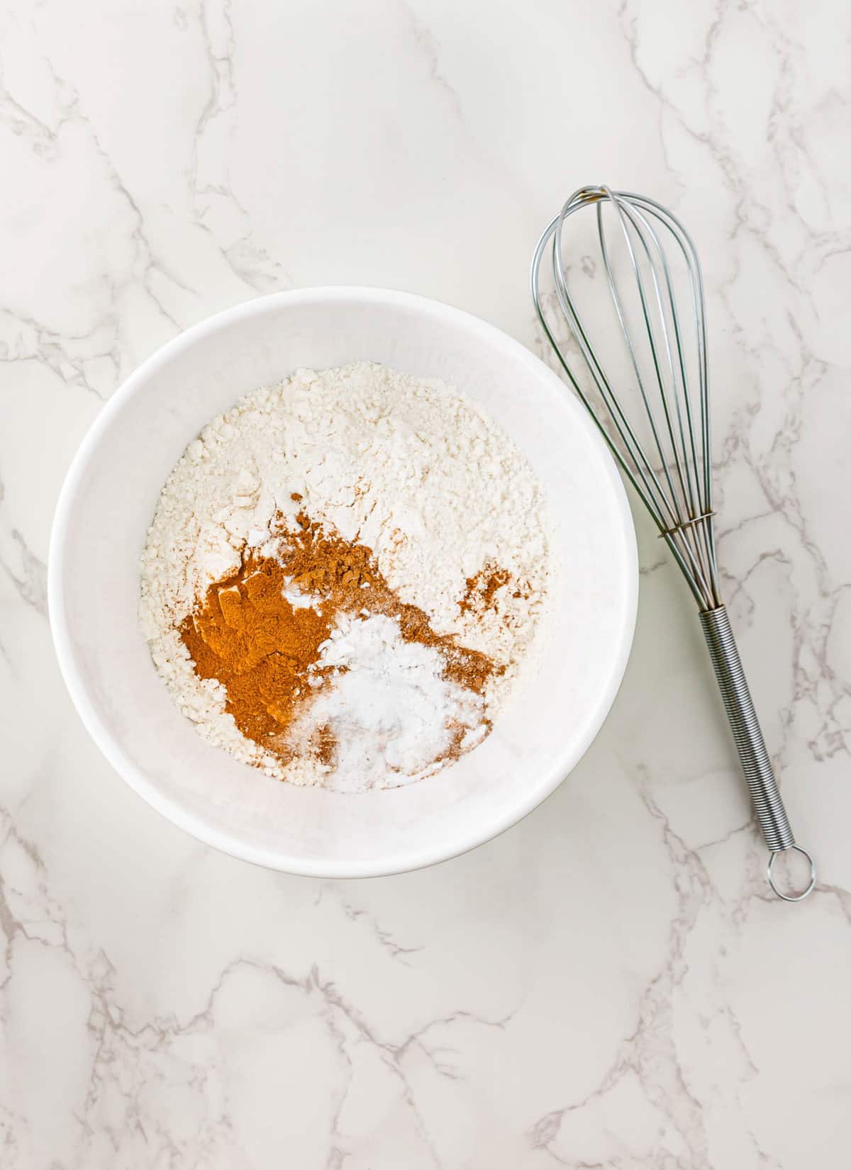 White bowl with flour, cinnamon, and baking soda&mdash;ready for a surprise Halloween treat like Pumpkin Banana Bread&mdash;next to a metal whisk on a marble countertop.