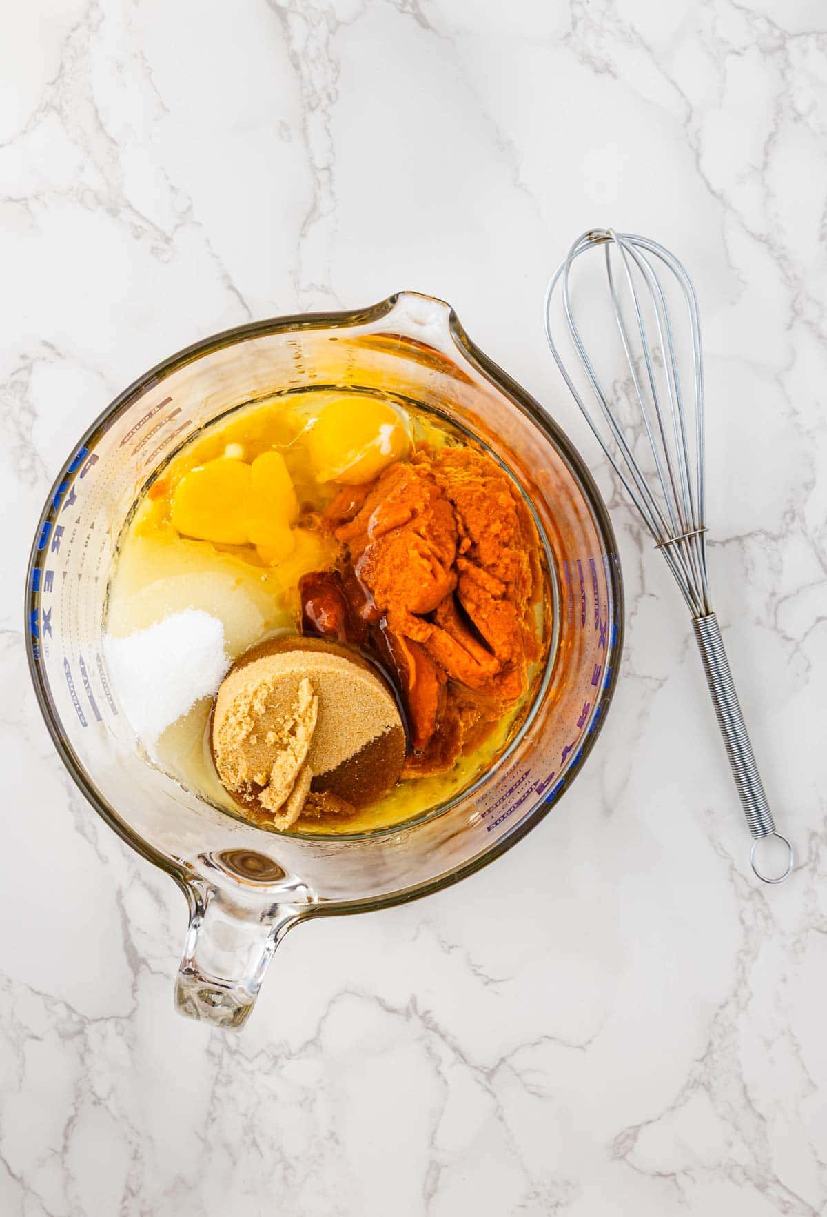 Glass bowl with eggs, canned pumpkin, sugar, and brown sugar next to a metal whisk on a marble surface&mdash;perfect ingredients for making Pumpkin Banana Bread this Halloween.
