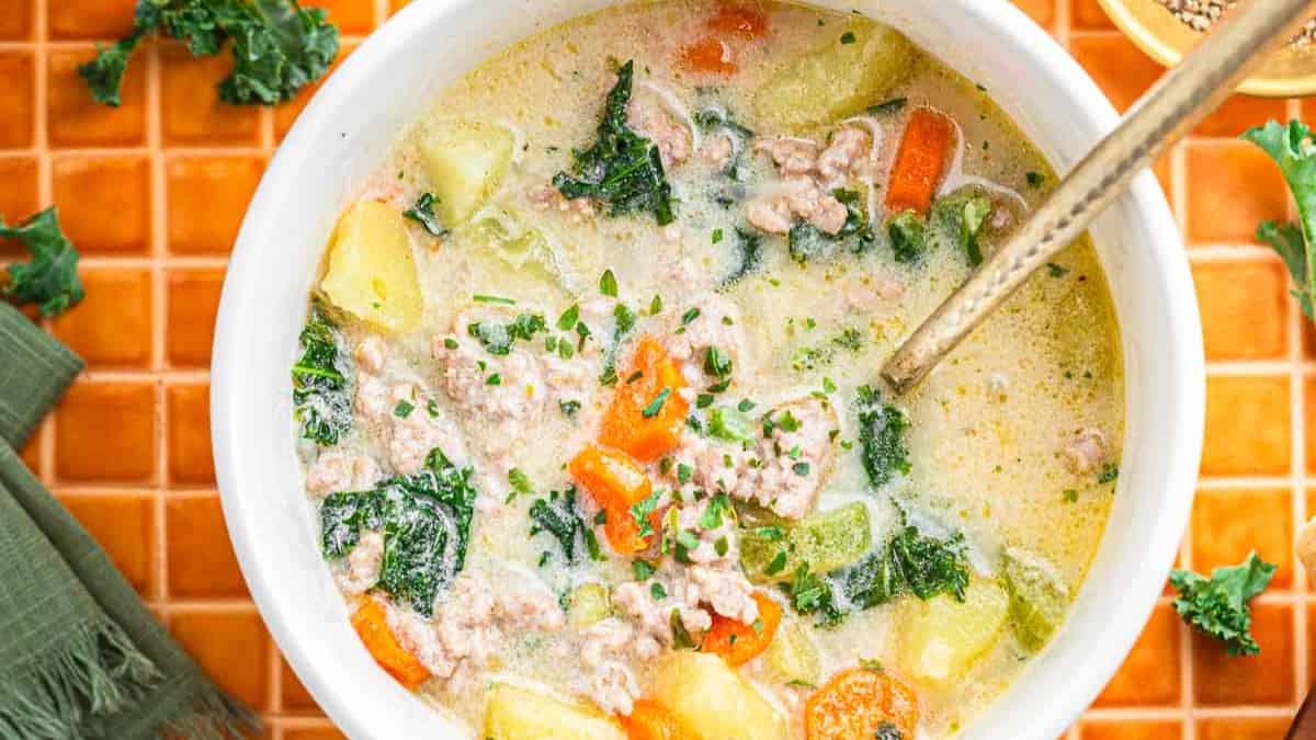 A bowl of creamy Tuscan Sausage and potato soup with ground meat, potatoes, carrots, kale, and herbs sits on an orange tiled surface.