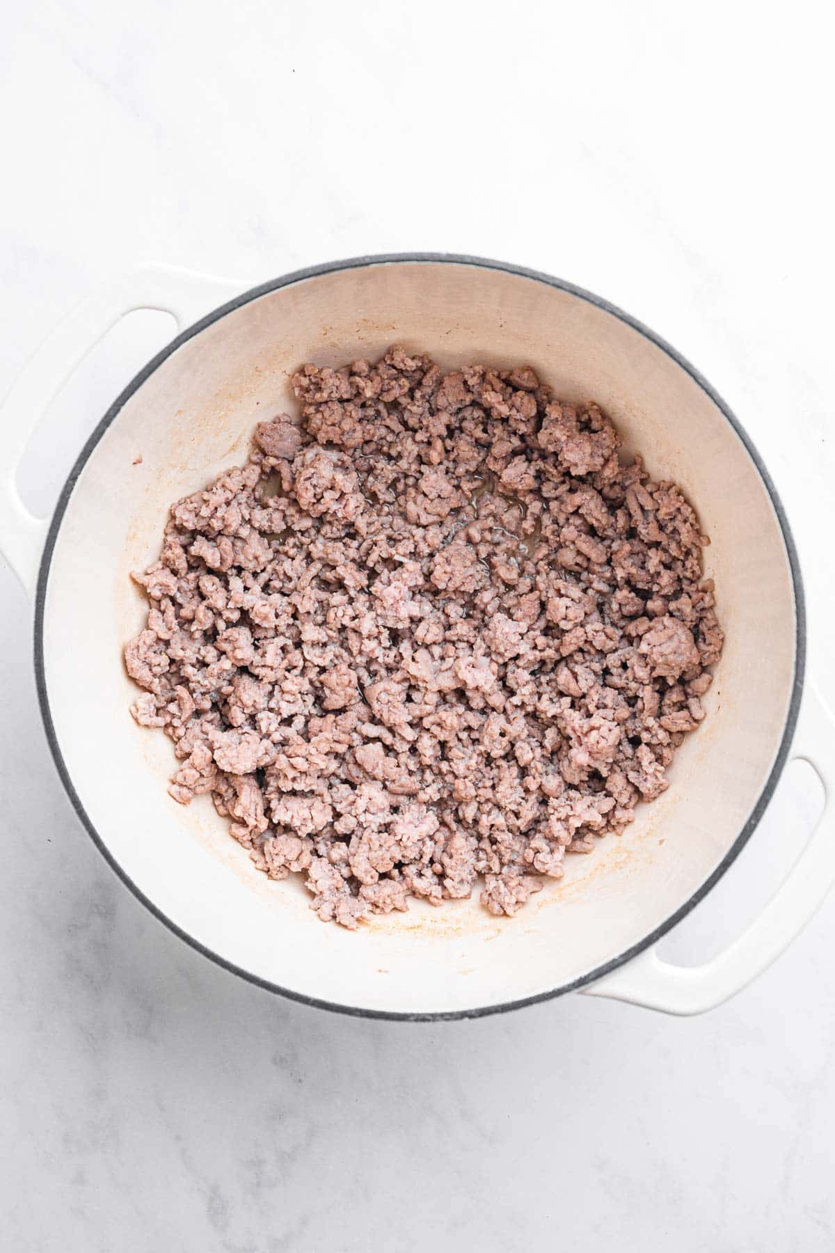 Cooked ground meat for a Tuscan Sausage recipe in a white pot on a marble countertop, viewed from above.