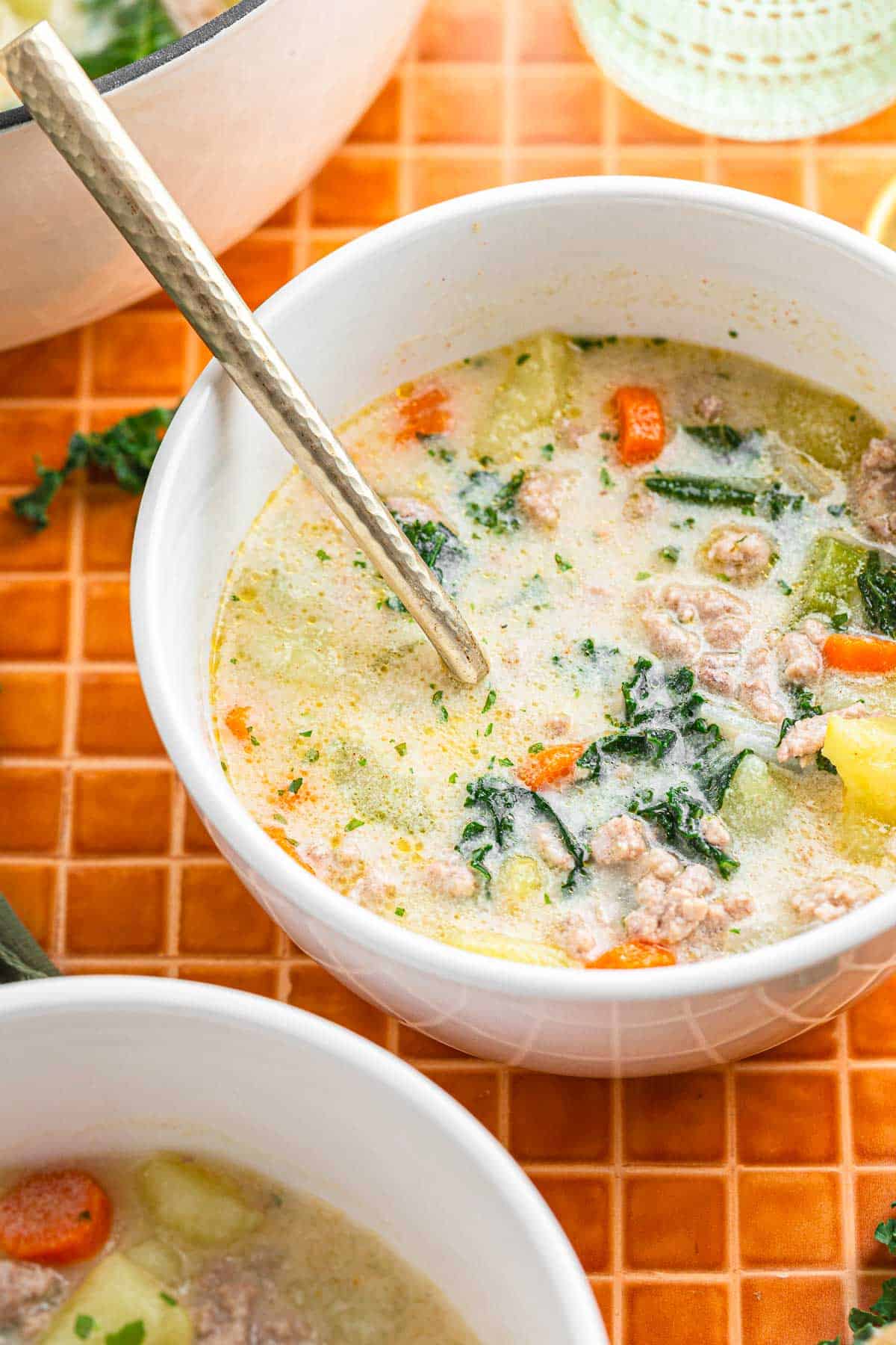 A bowl of creamy Tuscan sausage and potato soup with kale and carrots sits on an orange tiled surface.