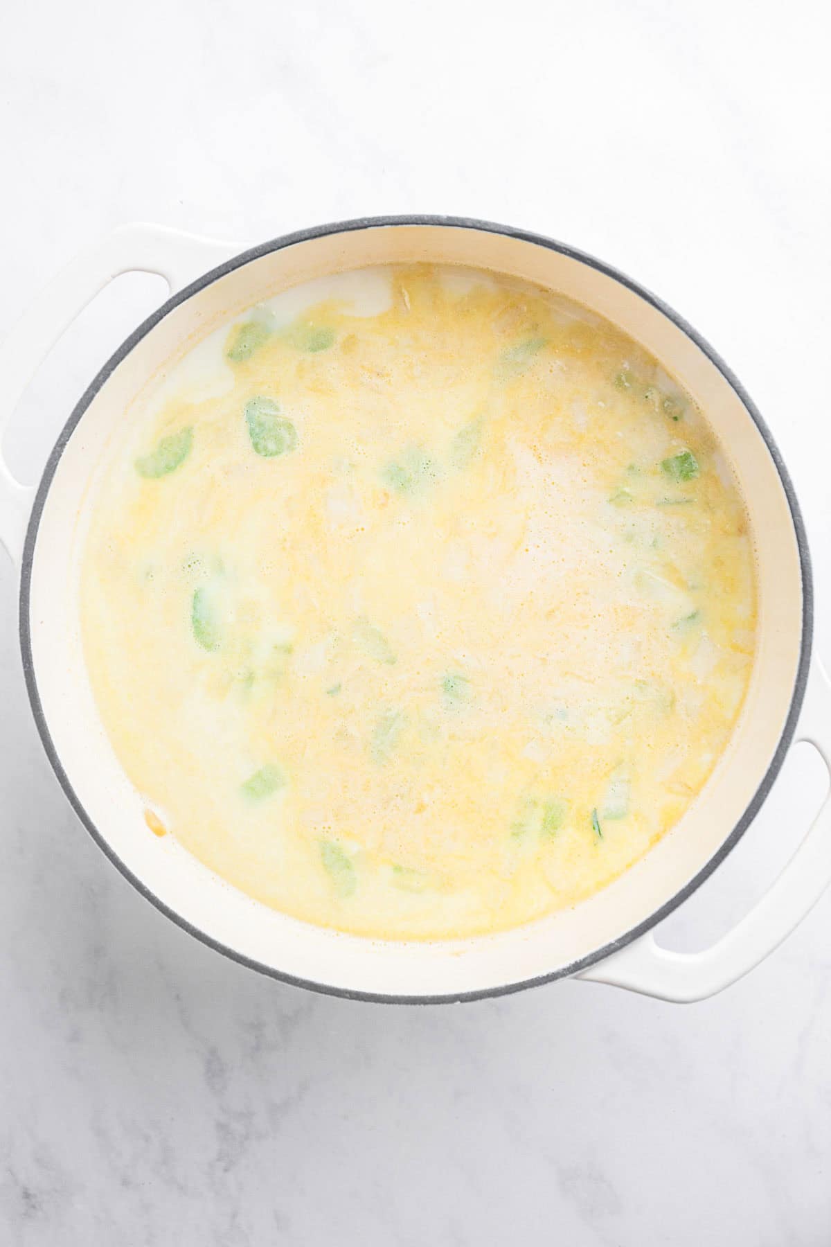 A white pot filled with creamy potato soup, featuring green vegetable pieces, sits on a white marble surface&mdash;a comforting recipe perfect for cozy nights.