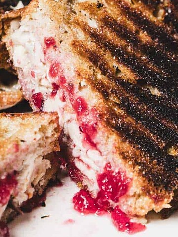 Close-up of a grilled sandwich with melted cheese, cranberry sauce, and herbs on toasted bread.