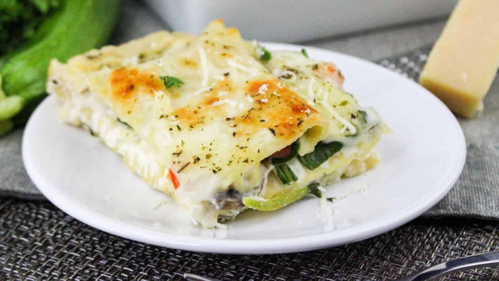 A slice of vegetable lasagna on a white plate, featuring layers of pasta, cheese, and vegetables, garnished with herbs and grated cheese.