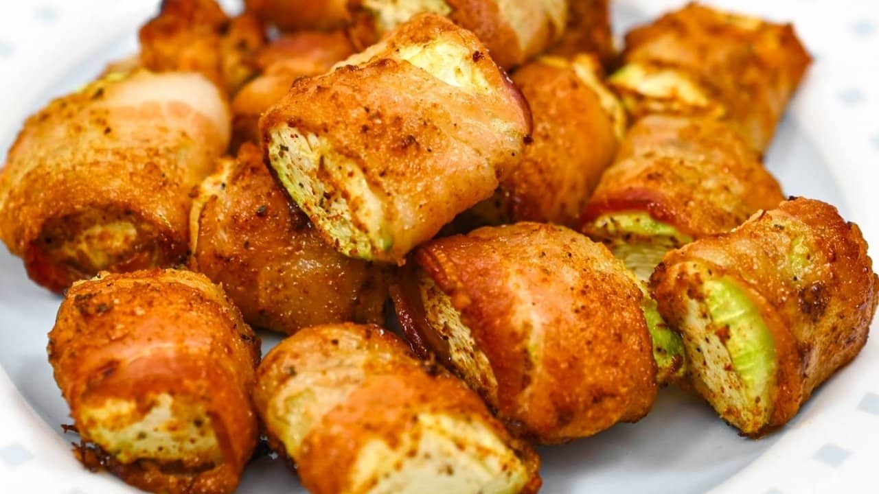 A plate of bacon-wrapped zucchini pieces, seasoned and baked, arranged closely together on a white dish.