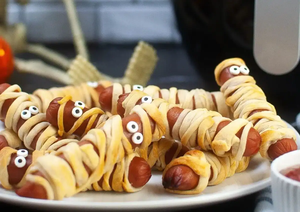 Hot dogs wrapped in dough to look like mummies, with candy eyes, arranged on a white plate for Halloween.