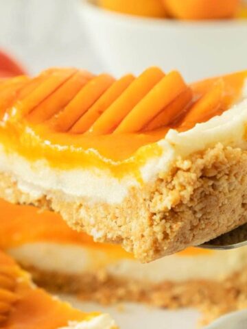 A slice of mango cheesecake with a crumbly crust and fresh mango slices on top, held up on a serving spatula.