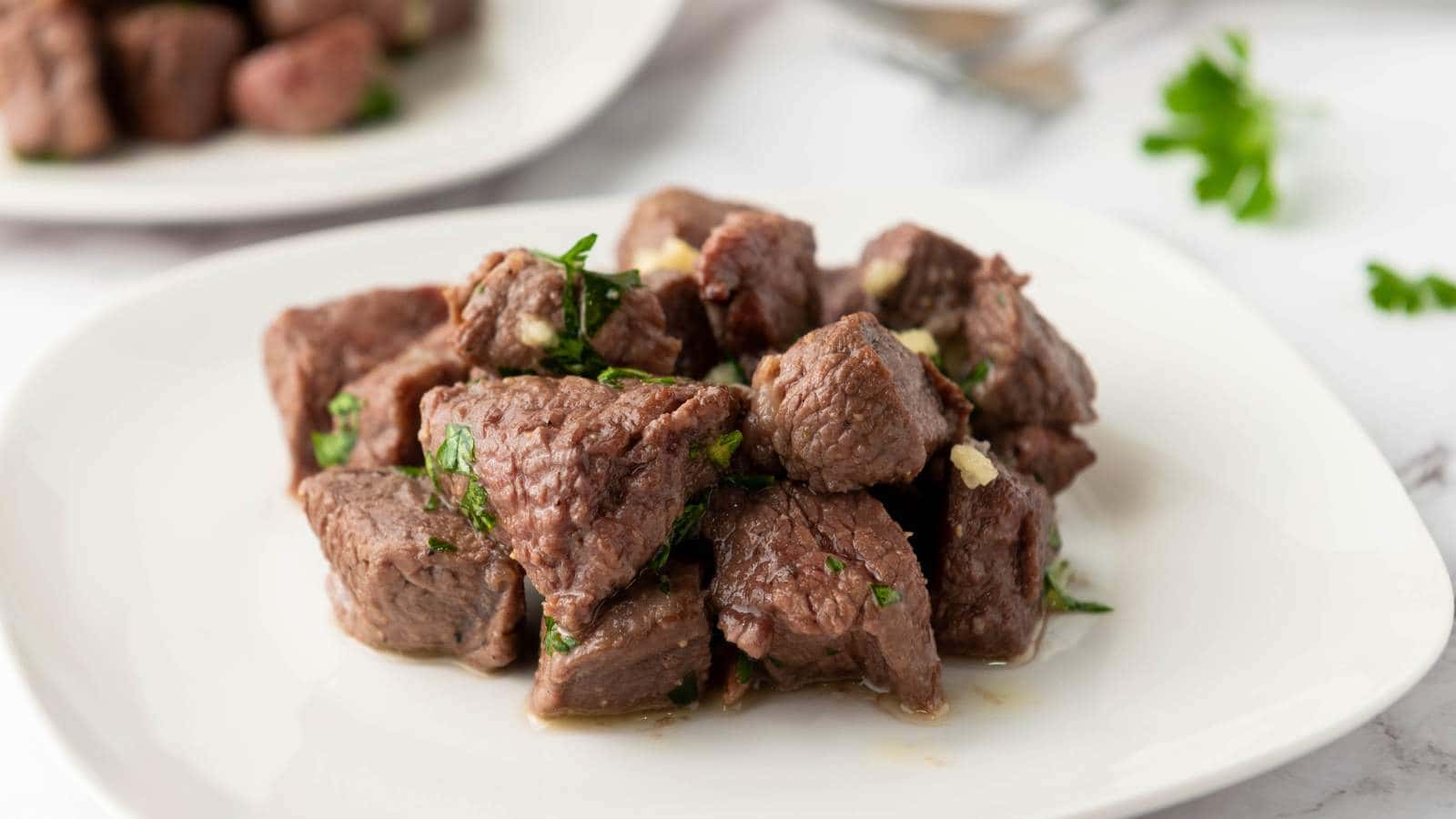A white plate with cooked steak bites garnished with chopped herbs.